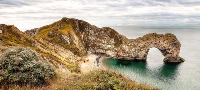 Jurassic Coast Eine Gruppe von Menschen steht an einem Strand in der Nähe eines Felsenbogens im Ozean