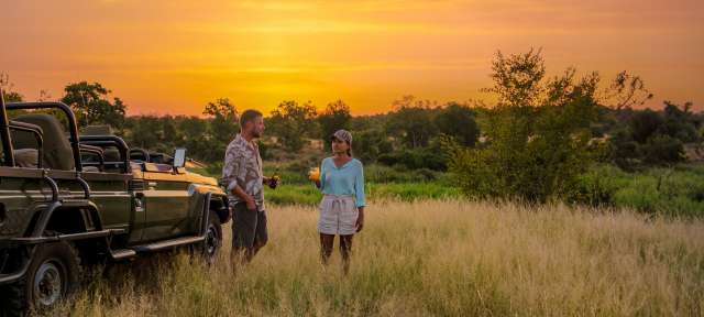 Sundowner nach einer Safari: Südafrika Ein Mann und eine Frau stehen in einem Feld vor einem Jeep