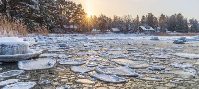 Wintersonne über dem eisbedeckten Meer vor dem estnischen Fischerdorf Altja mit verschneiten Holzhäusern und Bäumen.