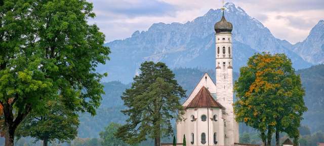 Eine kleine weiße Kirche steht inmitten eines grünen Feldes mit Bäumen und Bergen im Hintergrund