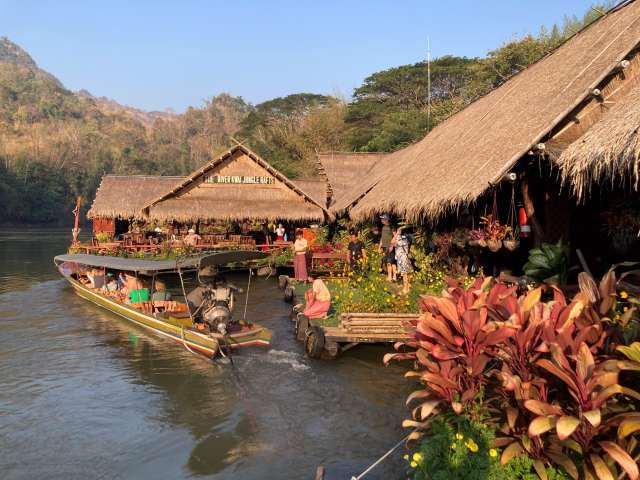 Ein Boot fährt an einem Restaurant mit Strohdach, das am Fluss Kwai liegt, vorbei.
