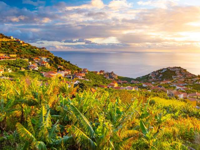 Ein Bergdorf mit roten Dächern, umgeben von Bananenplantagen, liegt am Meer unter einem dramatischen Himmel bei Sonnenuntergang.