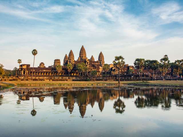 Die spiegelnde Wasseroberfläche vor Angkor Wat reflektiert die Türme des Tempels und die Palmen im goldenen Licht der Abendsonne.