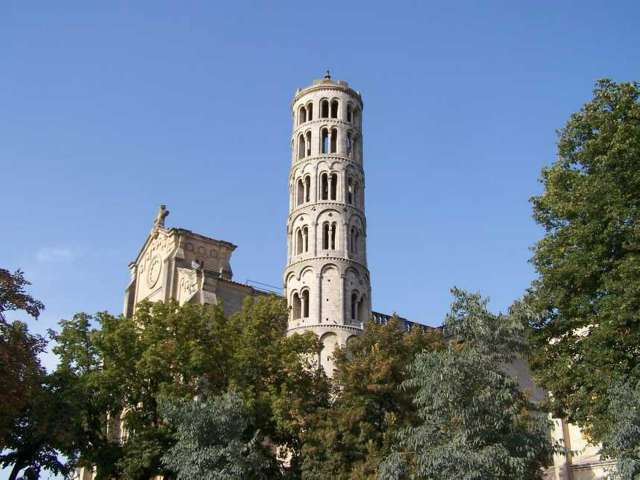 Eine romanische Kathedrale mit einem hohen, runden Glockenturm ragt im Hintergrund gegen einen klaren blauen Himmel über Bäumen auf.