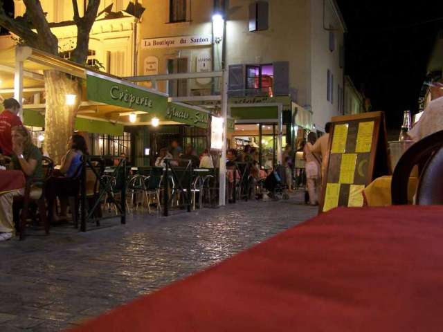 Nachts ist ein belebtes Straßencafé in einer engen Gasse mit vielen Gästen zu sehen, die an Tischen sitzen, während im Hintergrund Gebäude beleuchtet sind.