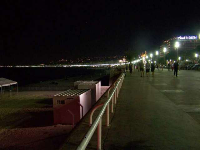 Am Abend spazieren Menschen auf einer beleuchteten Promenade entlang der Küste, hinter der sich eine Stadt mit vielen Lichtern erstreckt.