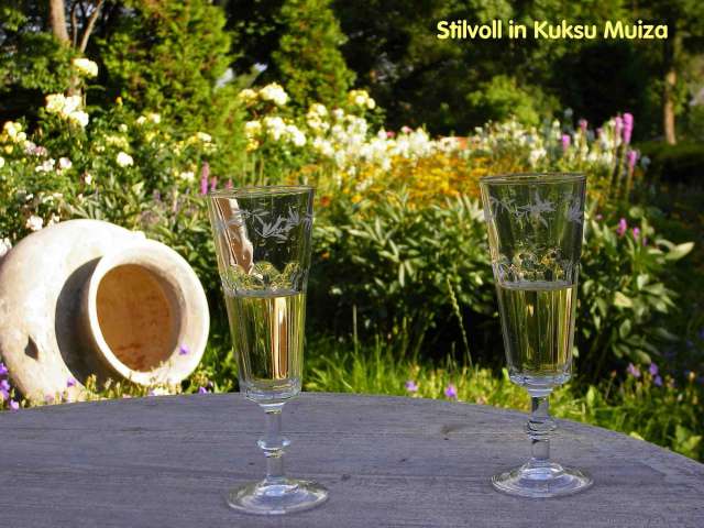 Zwei Weingläser mit goldenem Wein stehen auf einem Holztisch vor einem blühenden Garten.