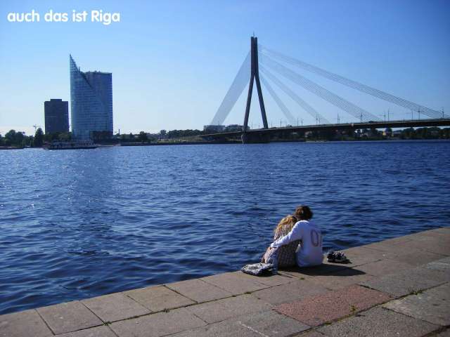Ein Paar sitzt am Ufer eines Flusses und blickt auf die Skyline von Riga, mit einer Brücke und Hochhäusern im Hintergrund.