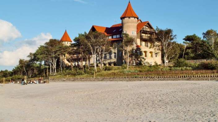Ein großes Gebäude mit roten Dächern sitzt auf einem Hügel über einem Strand