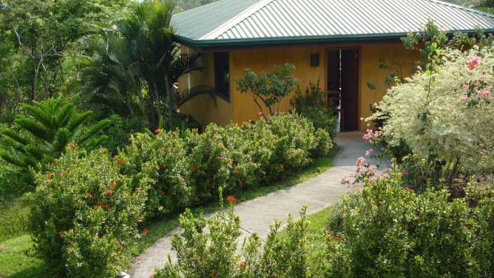 Ein gelbes Haus mit einem grünen Dach umgeben von Bäumen und Blumen