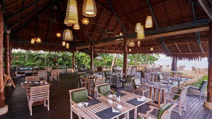 Ein rustikales Restaurant am Strand mit vielen Holztischen und -stühlen unter einem strohgedeckten Dach.