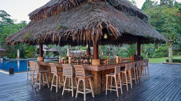 Ein überdachter Bar-Bereich im Freien mit hölzernen Barhockern steht am Rande eines Pools, umgeben von üppiger Vegetation.