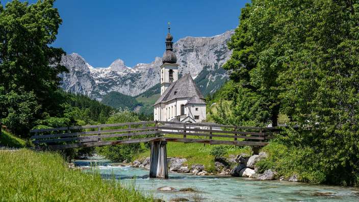 Eine Holzbücke überspannt einen klaren Fluss in den bayerischen Alpen, mit einer Kirche und schroffen Bergen im Hintergrund.