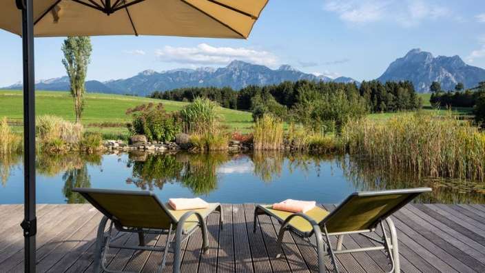 Ein Deck mit Liegestühlen und einem Regenschirm mit Blick auf einen Teich mit Bergen im Hintergrund