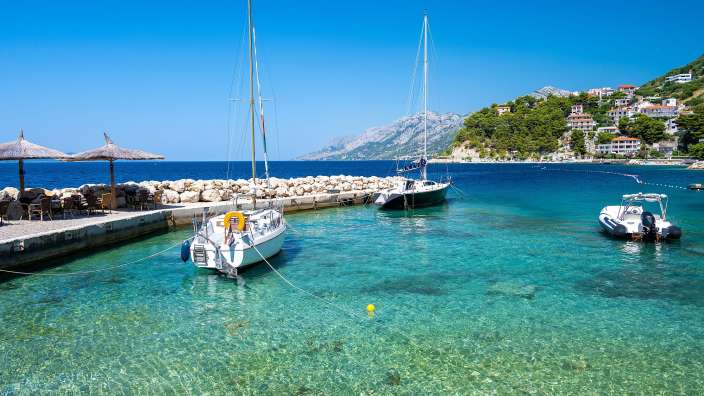 Ein Segelboot und ein Schlauchboot liegen in kristallklarem, türkisfarbenem Wasser vor einer Küste mit Häusern und Bergen.
