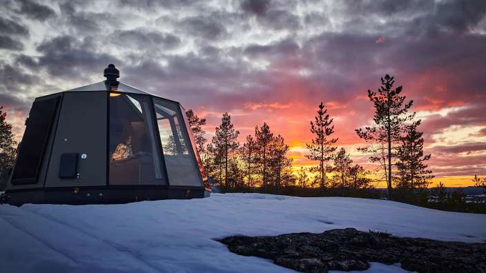 Ein Zelt sitzt auf einem schneebedeckten Hügel mit Bäumen im Hintergrund bei Sonnenuntergang