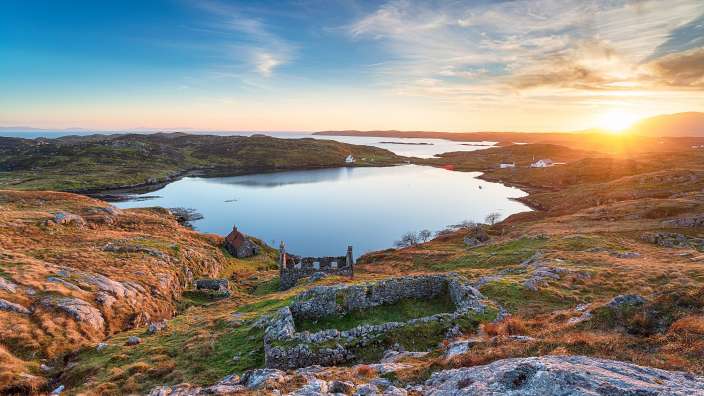 Eine malerische Aufnahme des Sonnenuntergangs über einer schottischen Küstenlandschaft mit zerklüfteten Hügeln, einem ruhigen See und den Ruinen alter Gebäude.