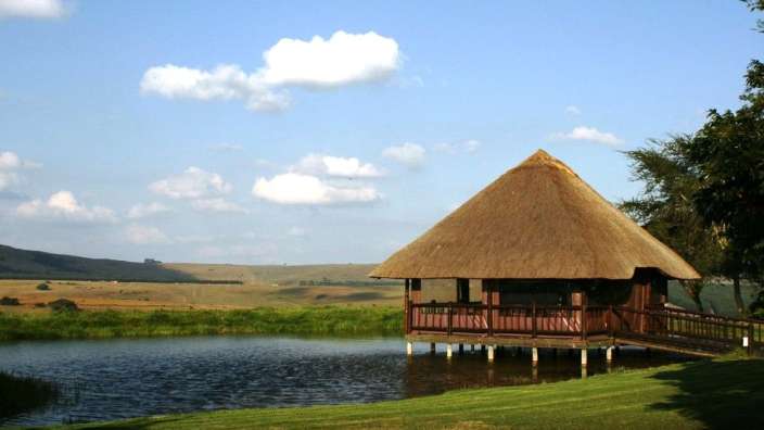 Ein Holzhaus mit Strohdach steht an einem Teich