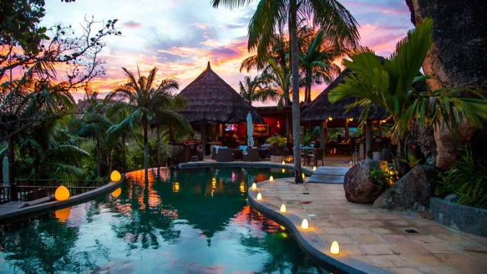 Ein großer Swimmingpool umgeben von Palmen mit einem Sonnenuntergang im Hintergrund