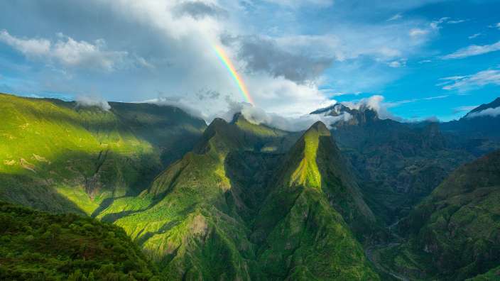 Ein Regenbogen erstreckt sich über grüne Berge und bewölkten Himmel.