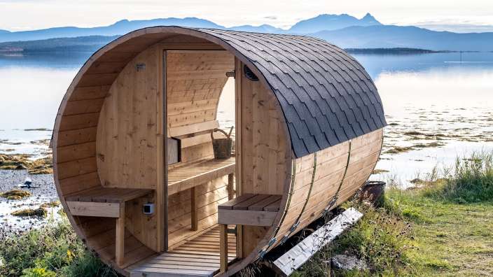 Eine Holzsauna mit einem schwarzen Dach sitzt am Ufer eines Sees