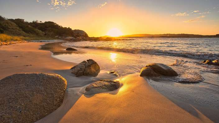 Ein sonniger Strand mit Felsen und dem Meer bei Sonnenuntergang.