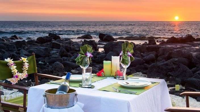 Ein Tisch mit zwei Stühlen und einem Eimer Champagner auf einem Strand bei Sonnenuntergang