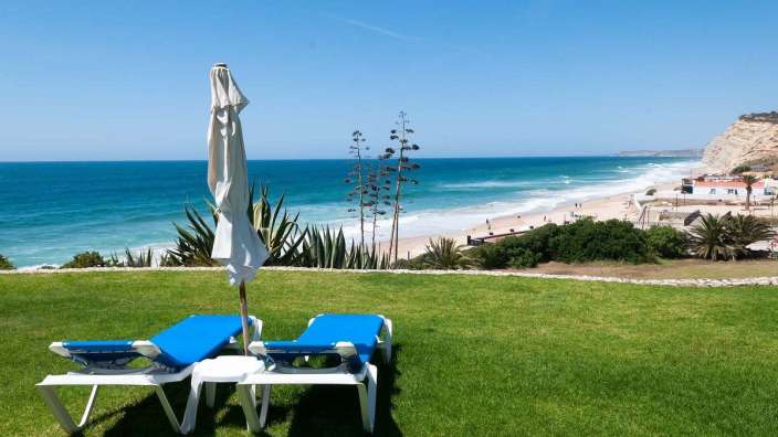 Zwei blaue Liegestühle und ein weißer Regenschirm auf einer Wiese mit Blick auf den Strand
