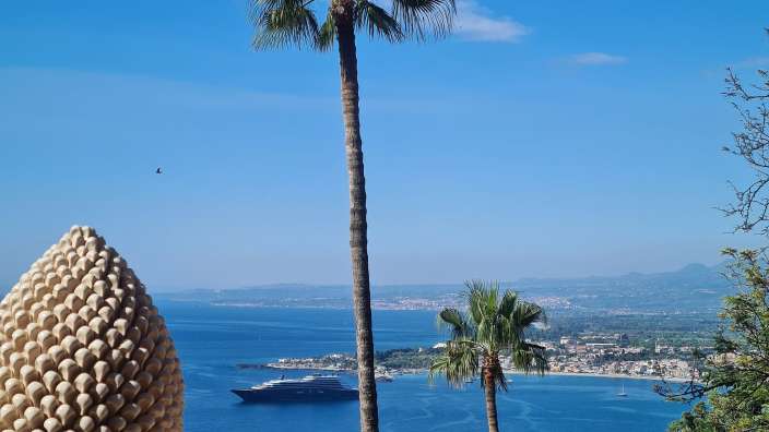 Taormina  Ein Blick auf das Meer mit Palmen und einer Tannenzapfenstatue im Vordergrund