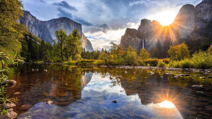 Die untergehende Sonne wirft einen warmen Schein auf die majestätischen Berge und Bäume, die sich im ruhigen Fluss spiegeln.