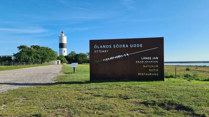 Ein braunes Schild steht in einem grasbewachsenen Feld neben einem Leuchtturm