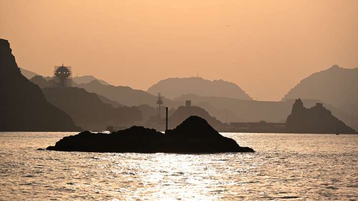 Silhouetten von Bergen, einer felsigen Insel und einer ungewöhnlichen Struktur mit Gerüsten vor einem sonnenuntergangsfarbenen Himmel über dem Meer.