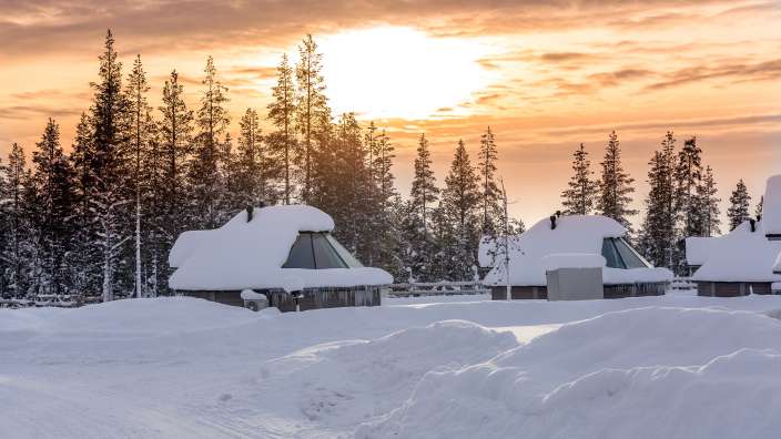 Ein verschneites Dorf mit kleinen Holzhäusern, die mit viel Schnee bedeckt sind, liegt vor einem Wald und einem Himmel mit orangefarbenem Sonnenuntergang.