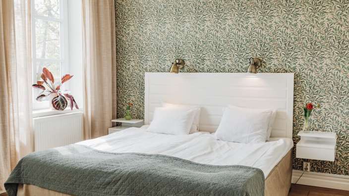 Ein Doppelbett mit weißer Bettwäsche und einer grünen Decke steht vor einer grünen Tapete mit Blattmuster, neben einem Fenster mit hellrosa Vorhängen.