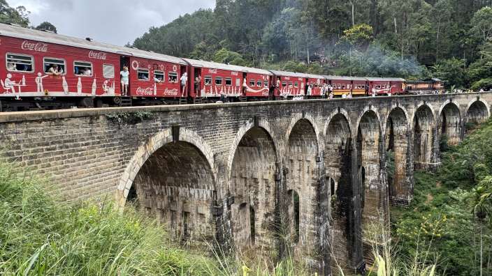 Ein roter Zug mit der Aufschrift "Coca-Cola" fährt auf einer gemauerten Bogenbrücke inmitten einer üppig grünen Landschaft.