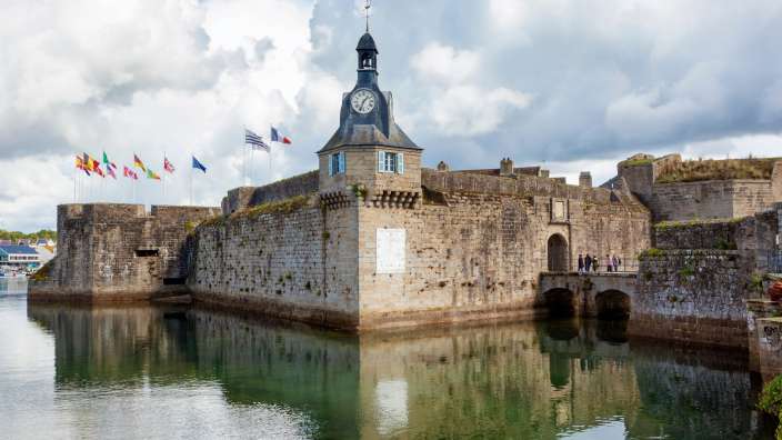 Eine historische Festung mit einem Uhrenturm und vielen Flaggen wird von Wasser umgeben, dessen Oberfläche die Gebäude und den Himmel widerspiegelt.