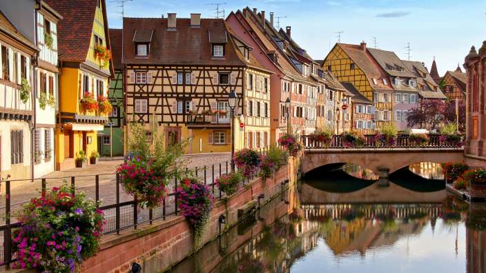 Das Bild zeigt eine Reihe von traditionellen Fachwerkhäusern entlang eines Kanals mit einer Brücke, die von üppigen Blumen und Pflanzen geschmückt ist.