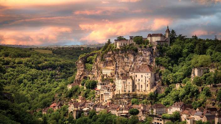 Eine malerische Ansicht eines mittelalterlichen Dorfes und seiner Burg, die an einer steilen Klippe in Frankreich im goldenen Licht des Sonnenuntergangs liegt.