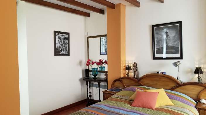 In einem Schlafzimmer mit Holzbalkendecke und orangefarbenen Säulen stehen ein Bett mit farbenfroher Bettwäsche und ein kleiner Nachttisch neben einem Spiegel.