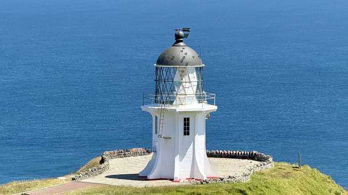 Ein weißer Leuchtturm steht auf einem grünen Hügel am Meer unter einem blauen Himmel.