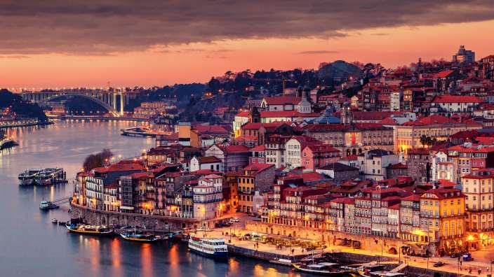 Eine malerische Aufnahme der Stadt Porto bei Sonnenuntergang, mit dem Douro-Fluss im Vordergrund und der charakteristischen Architektur im Hintergrund.