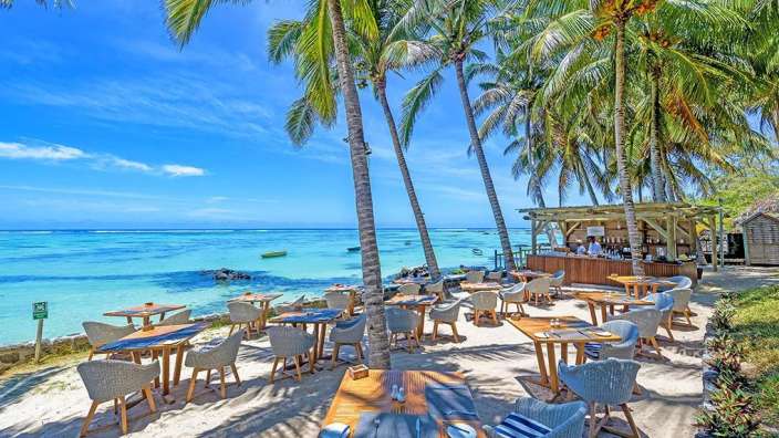 Ein tropisches Strandrestaurant mit Tischen und Stühlen am Meer unter Palmen an einem sonnigen Tag.