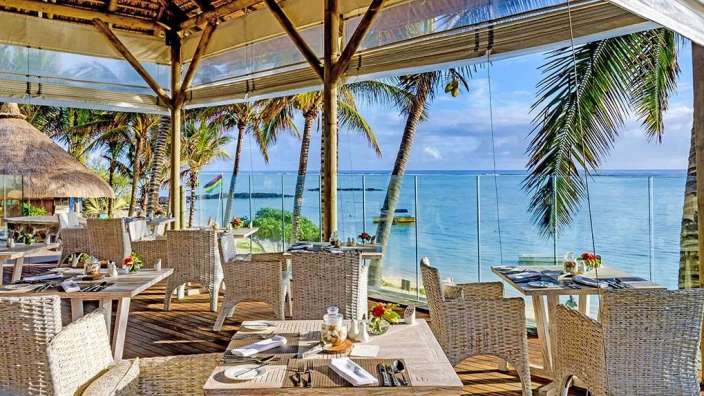 Ein Tisch ist in einem Restaurant mit Blick auf das Meer gedeckt, das von Palmen und einem tropischen Himmel umrahmt wird.