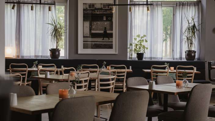Das Bild zeigt den Innenraum eines Restaurants mit vielen Holztischen und Stühlen, dekoriert mit Kerzen und kleinen Vasen mit Grünzeug.
