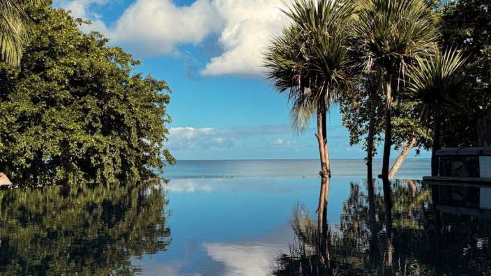 Ein Blick über einen Infinity-Pool auf das Meer, gesäumt von üppigem Grün und Palmen, bei strahlendem Sonnenschein und blauen Himmel mit Wolken.