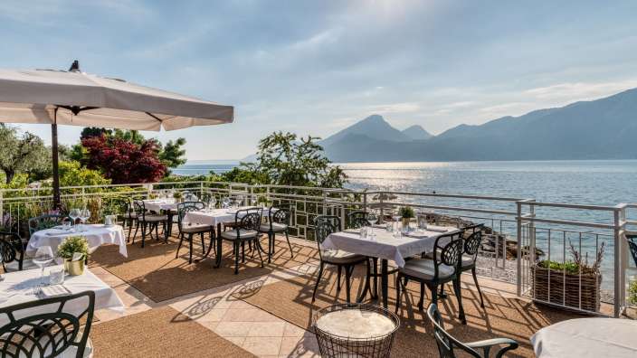 Auf einer Terrasse mit Blick auf das Meer und die Berge sind mehrere Tische für eine Mahlzeit gedeckt.