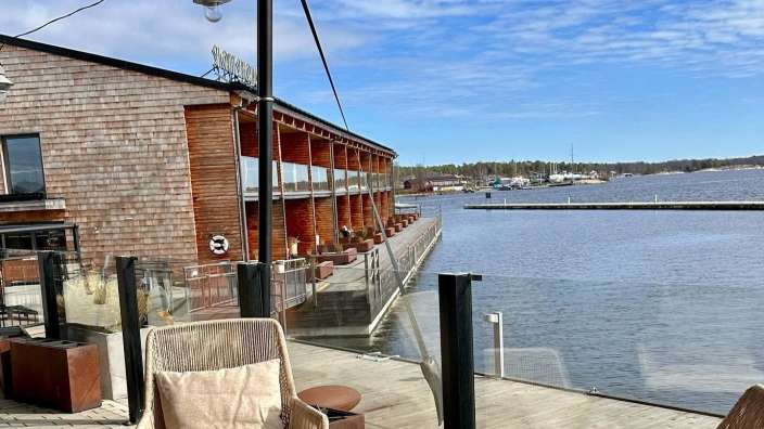 Auf dem Bild ist eine Terrasse mit Blick auf ein Gewässer mit Booten und einer Holzgebäudefassade zu sehen.