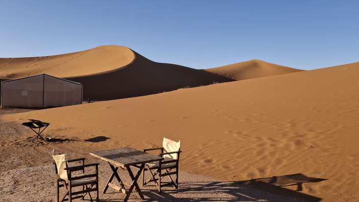 Ein Camp mit zwei Stühlen und einem Tisch steht am Rande von Sanddünen unter blauem Himmel.