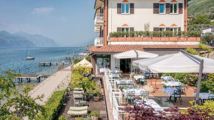 Ein Restaurant mit Blick auf einen See und Berge, im Vordergrund Tische und Stühle im Freien.
