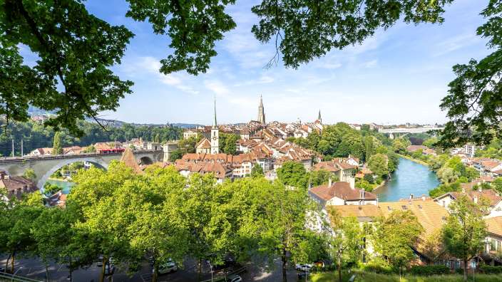 Eine Stadt mit roten Dächern und einer Brücke mit einer roten Bahn über einen blauen Fluss, umgeben von Bäumen, unter einem klaren Himmel.
